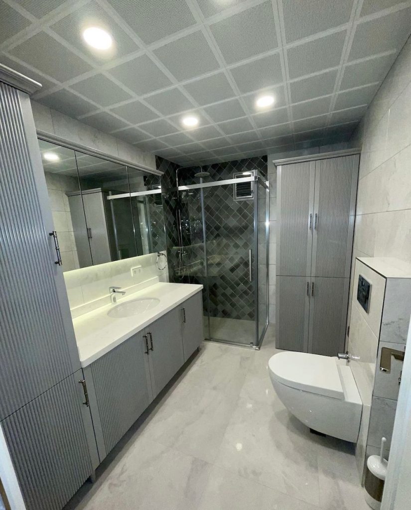 Gri çizgili kapaklı lavabo dolabı, geniş ayna, cam bölmeli duş alanı ve kare panelli asma tavanda spot aydınlatmalar bulunan banyo