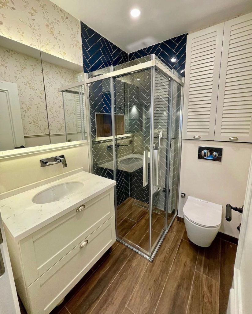 Lacivert balıksırtı fayanslı duş alanında cam duşakabin, asma klozet, aydınlatmalı ayna ve beyaz lavabo dolabı bulunan banyo